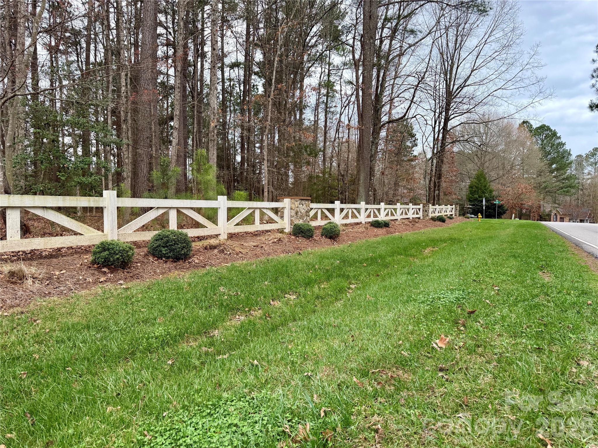 Property frontage with a white cross-braced fence, stone pillar, green lawn, and woods beside the road.