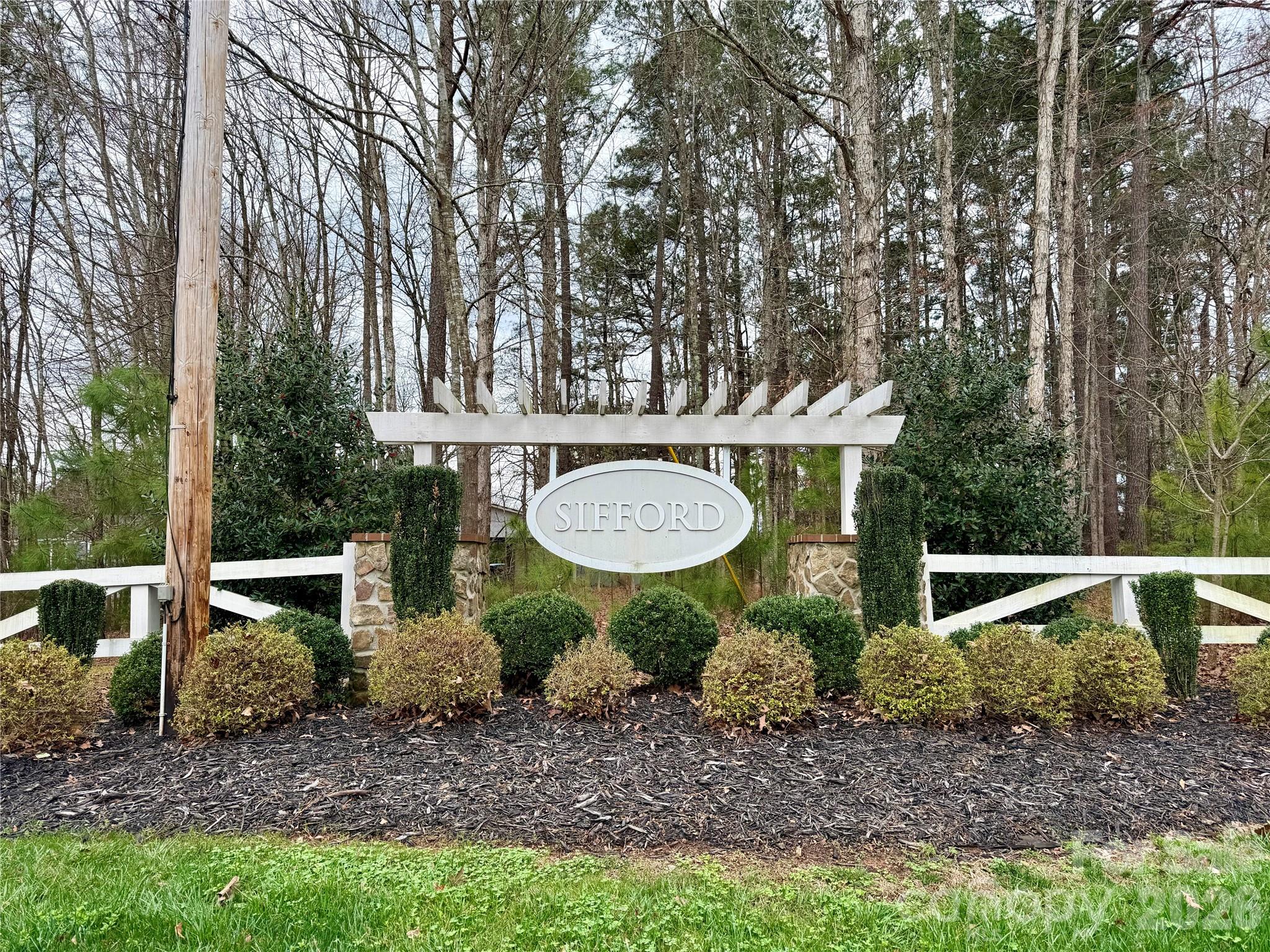 Sifford neighborhood entrance sign beneath a white pergola, surrounded by shrubs and white fencing.
