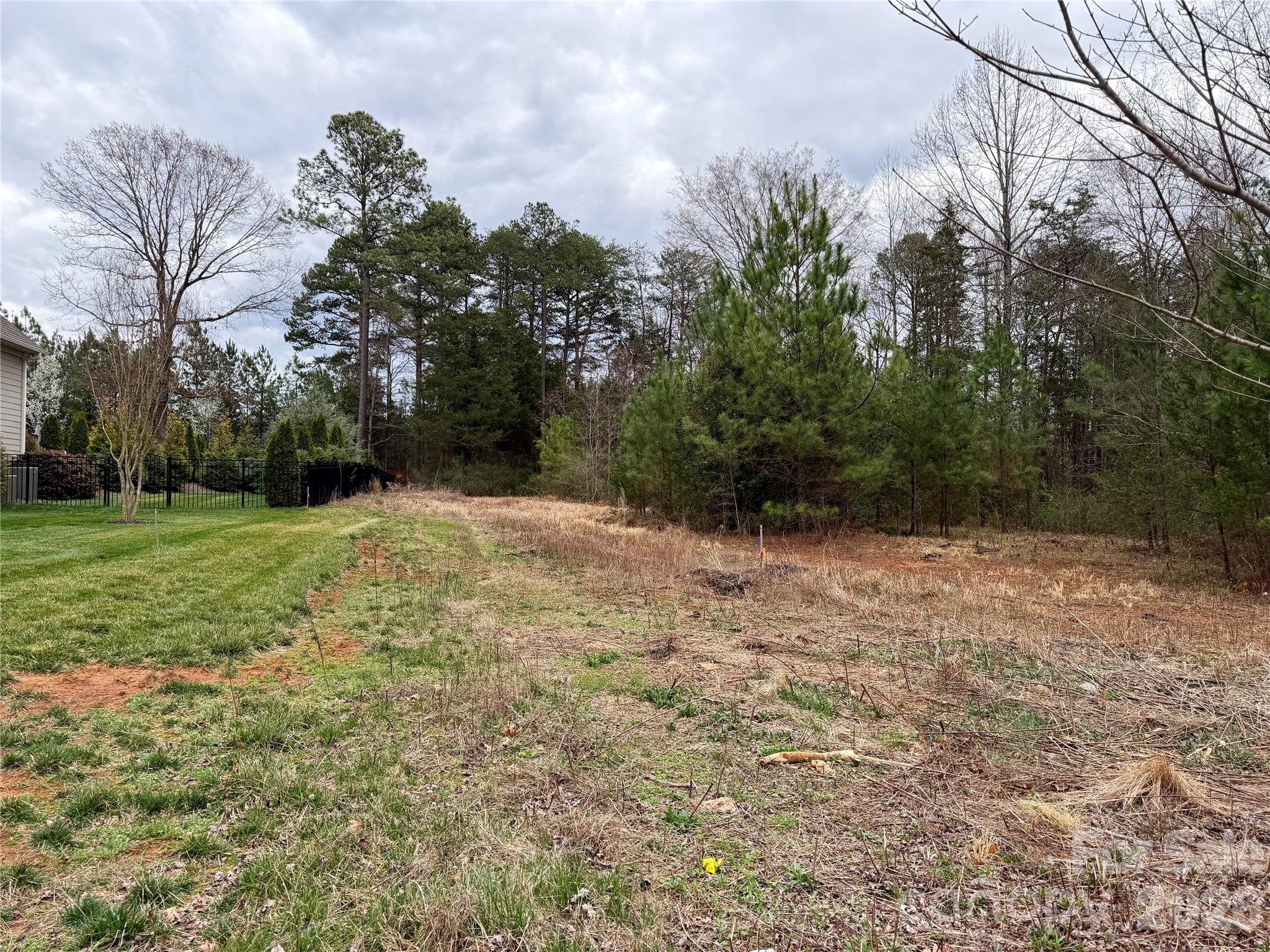 Vacant lot with dry brush, mature trees, and a neighboring home beside a black fence.