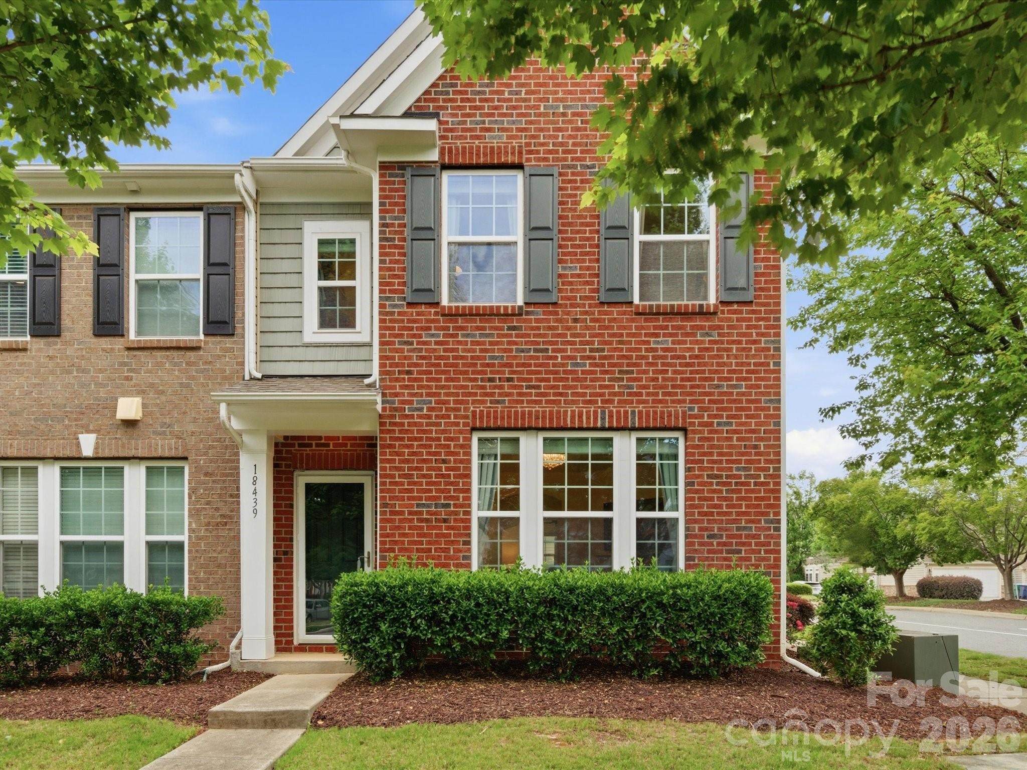 Two-story brick home with gray siding, dark shutters, and trimmed hedges at the front entry.