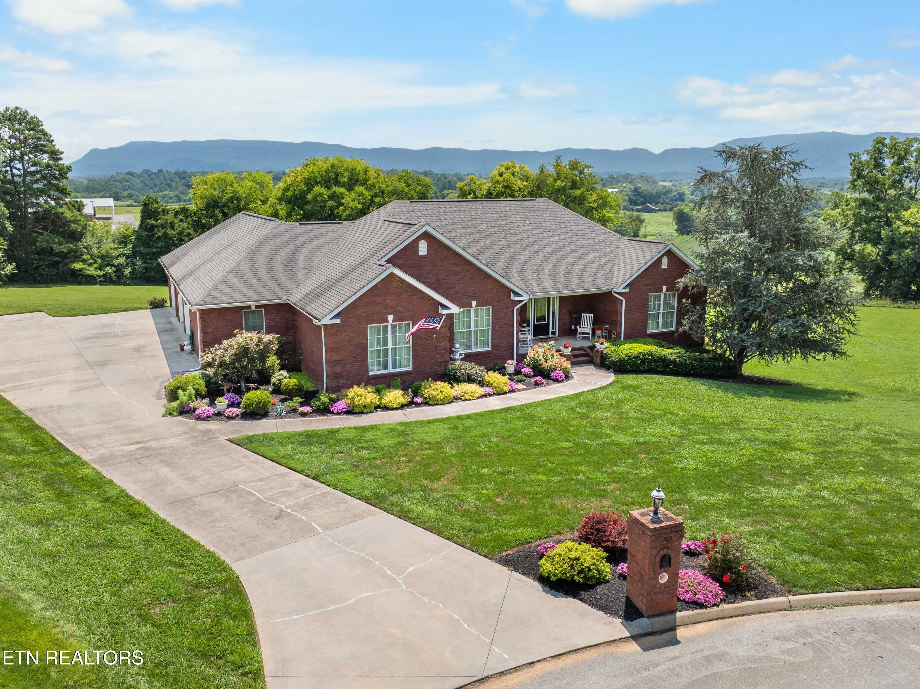Red-brick home with a curved driveway, colorful flower beds, and mountain views.