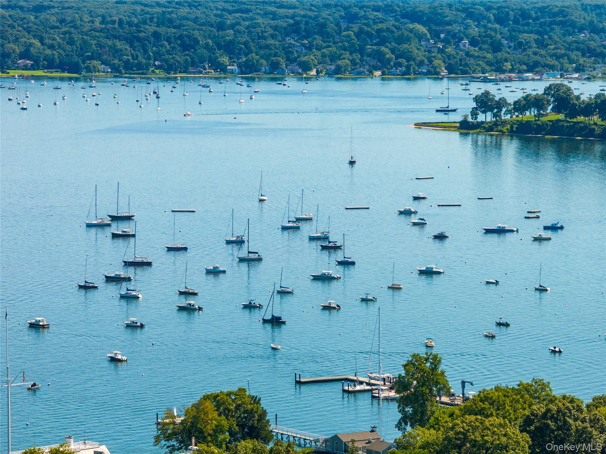 Harbor view dotted with sailboats and motorboats, with a wooded shoreline and dock.