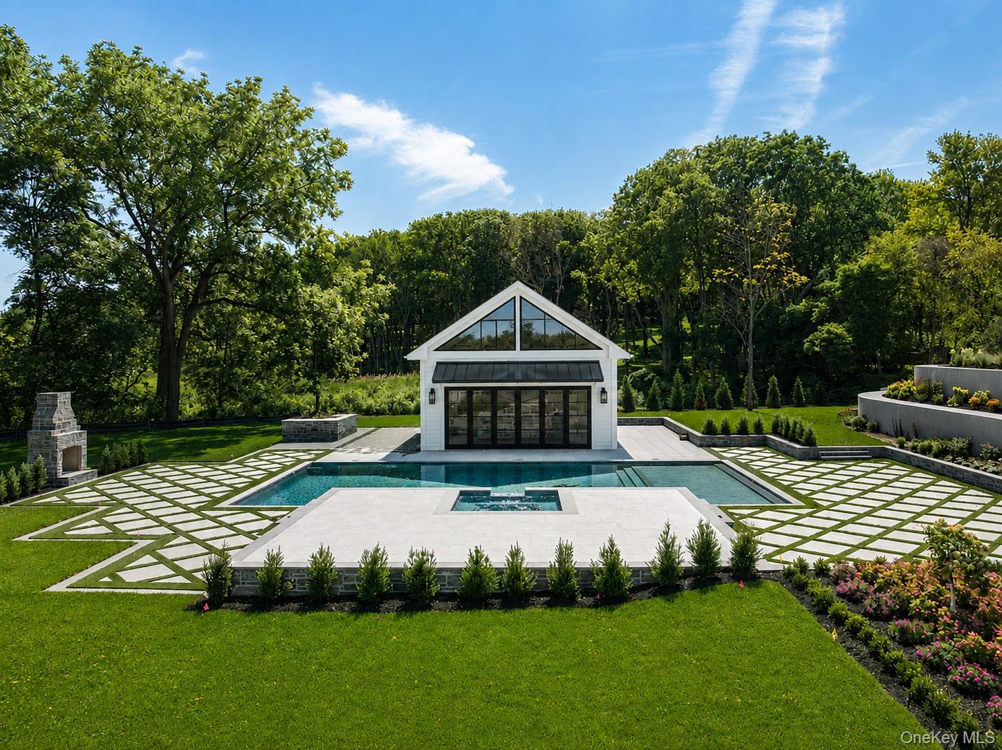 Backyard pool and raised spa in front of a white, glass-fronted pool house, surrounded by stone paths and trees.