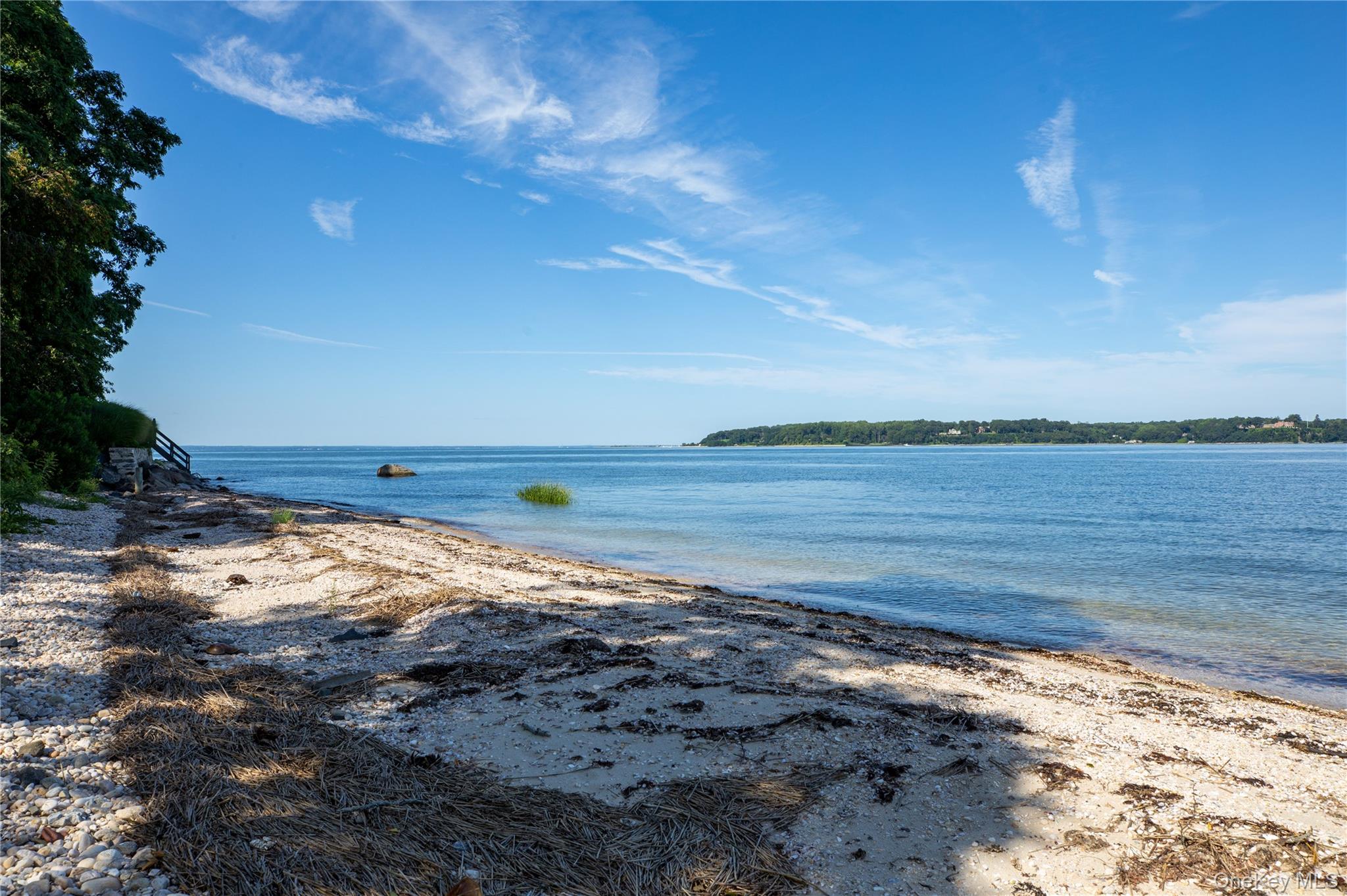 Pebble beach along calm blue water, with a wooded shoreline across the bay.