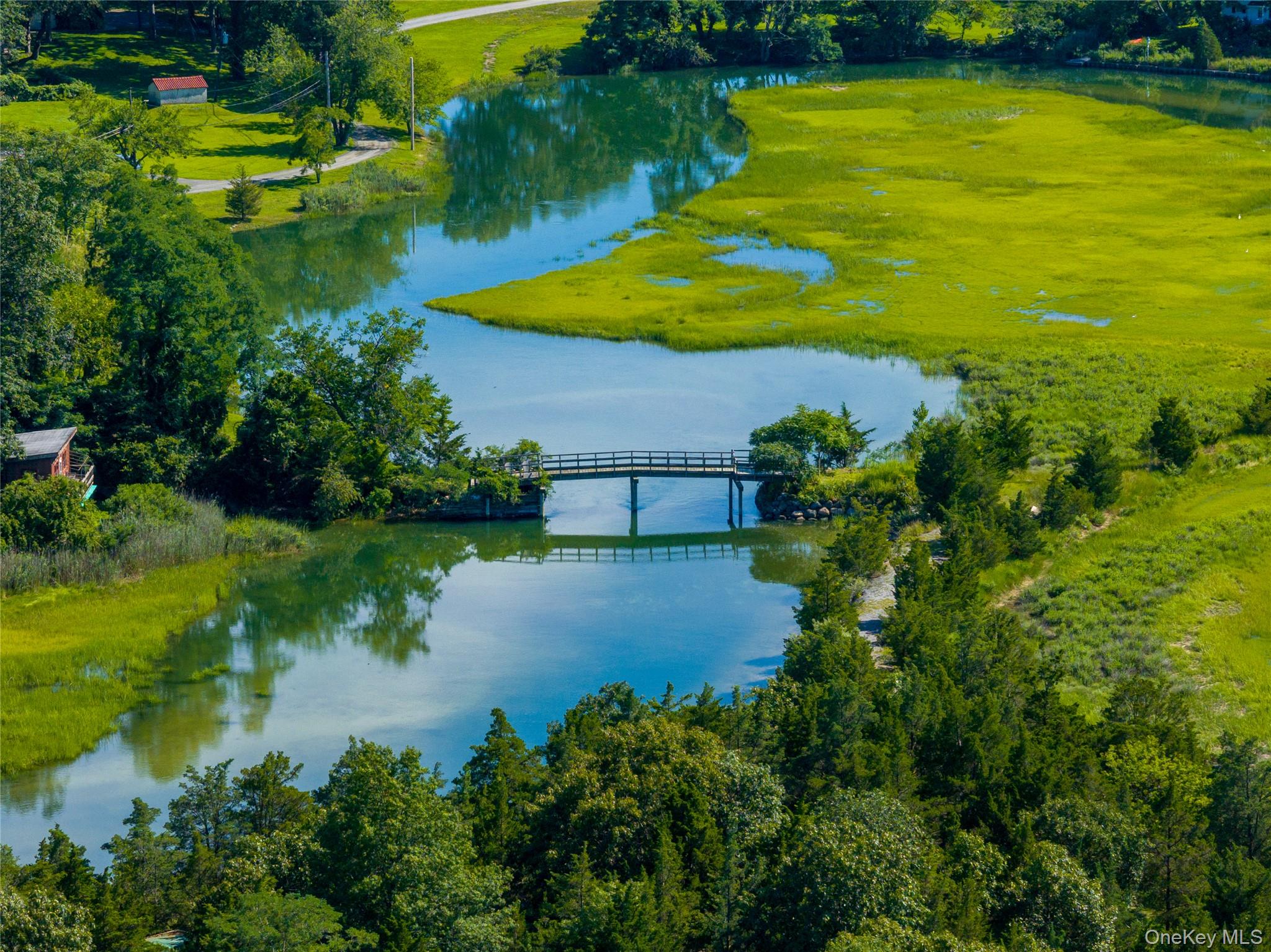 Aerial creek and marshland with a small bridge and wooded banks.