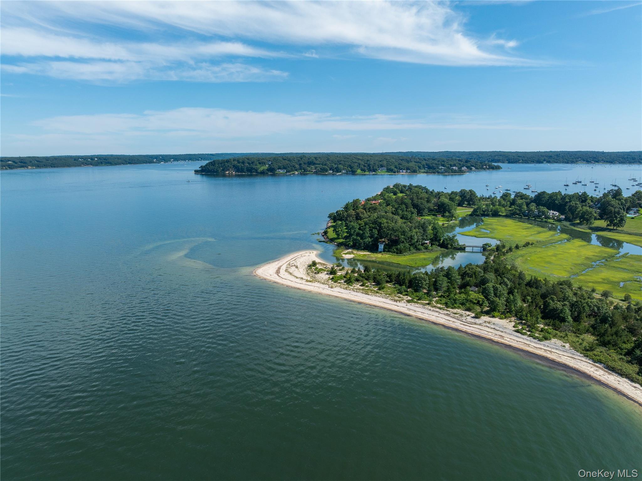 Aerial view of a wooded shoreline with a sandy beach, marshland, and sailboats across the bay.
