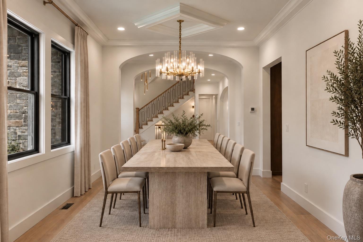 Dining room with a long stone table, modern chandelier, and staircase beyond an arched opening.