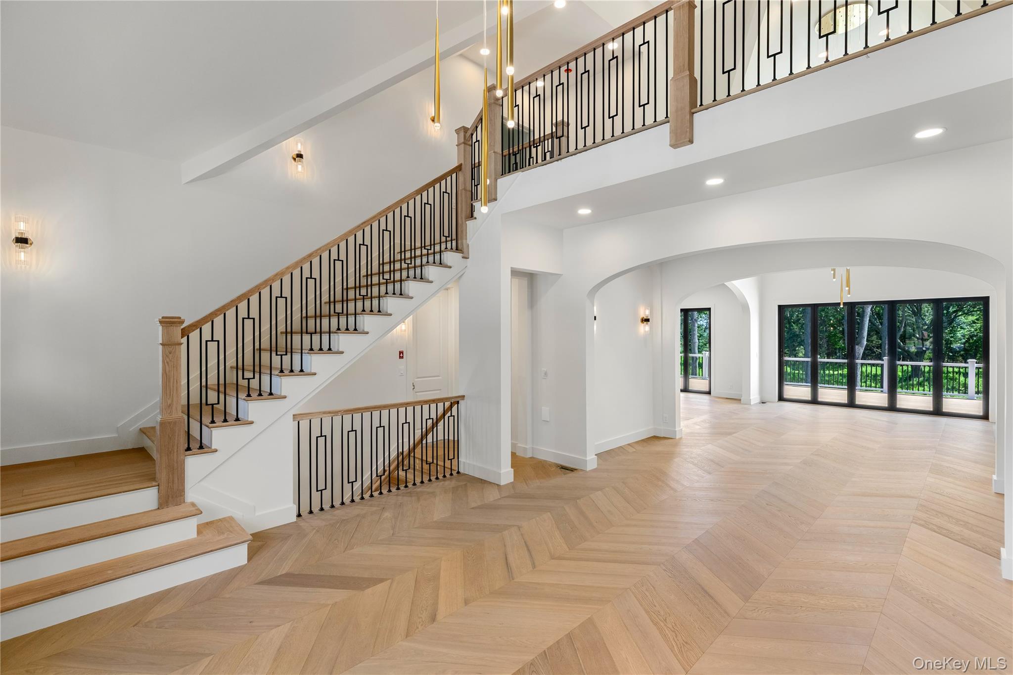 Two-story foyer with a wood staircase, geometric black railings, herringbone floors, and glass doors overlooking trees.