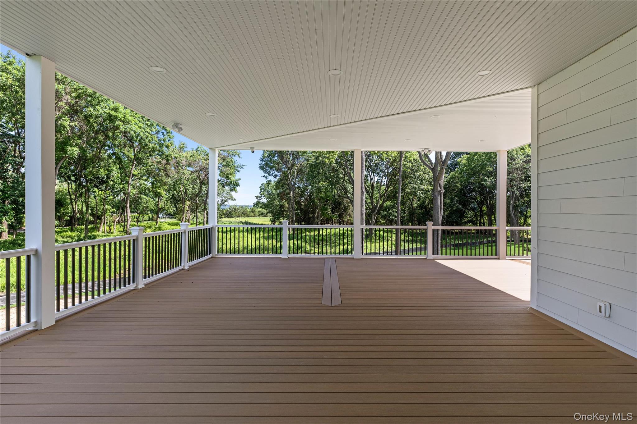 Covered deck with brown plank flooring and white railings overlooking a lawn and mature trees.