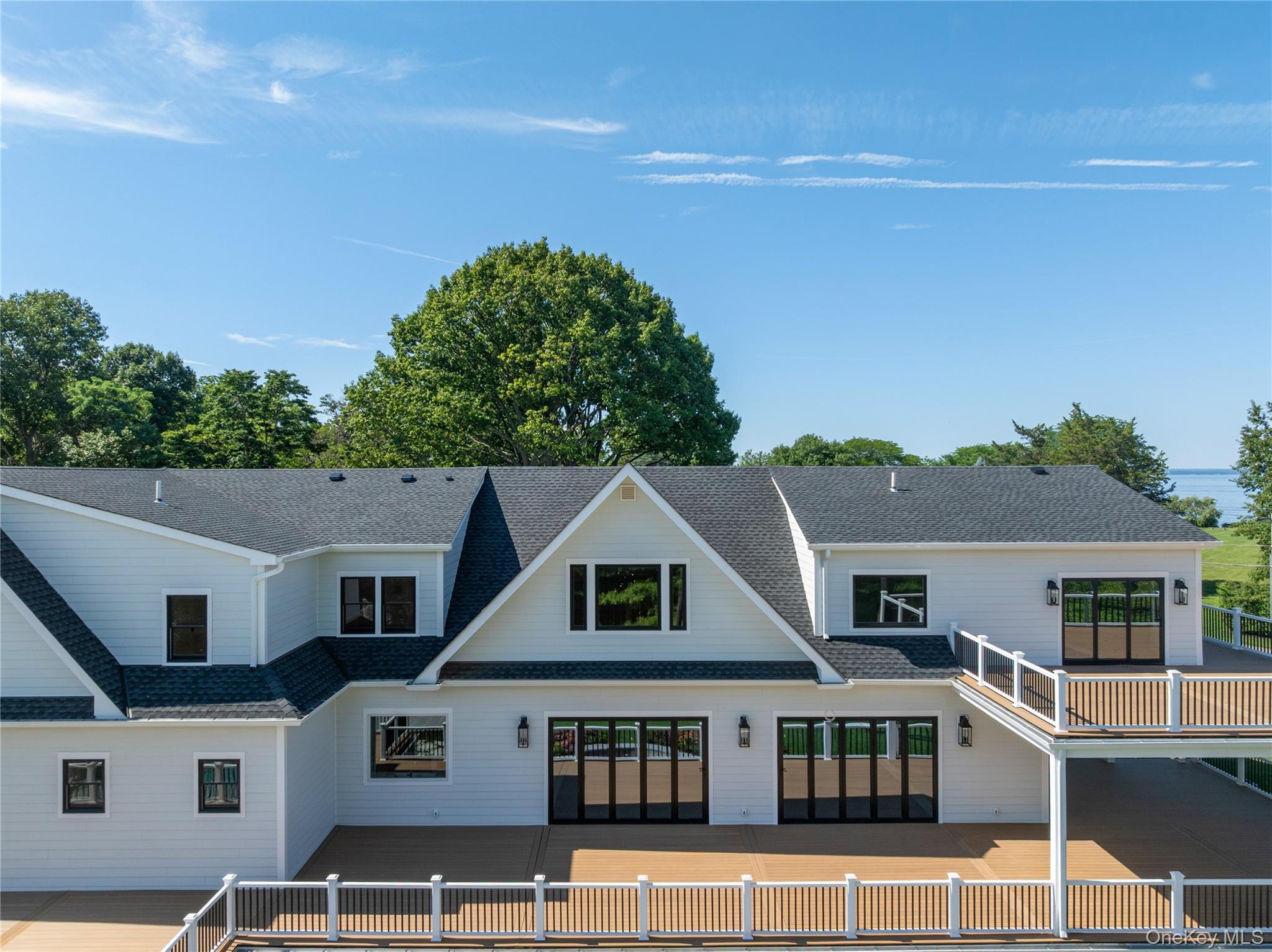 White gabled house with expansive wood decks and a raised balcony, with water visible beyond the trees.