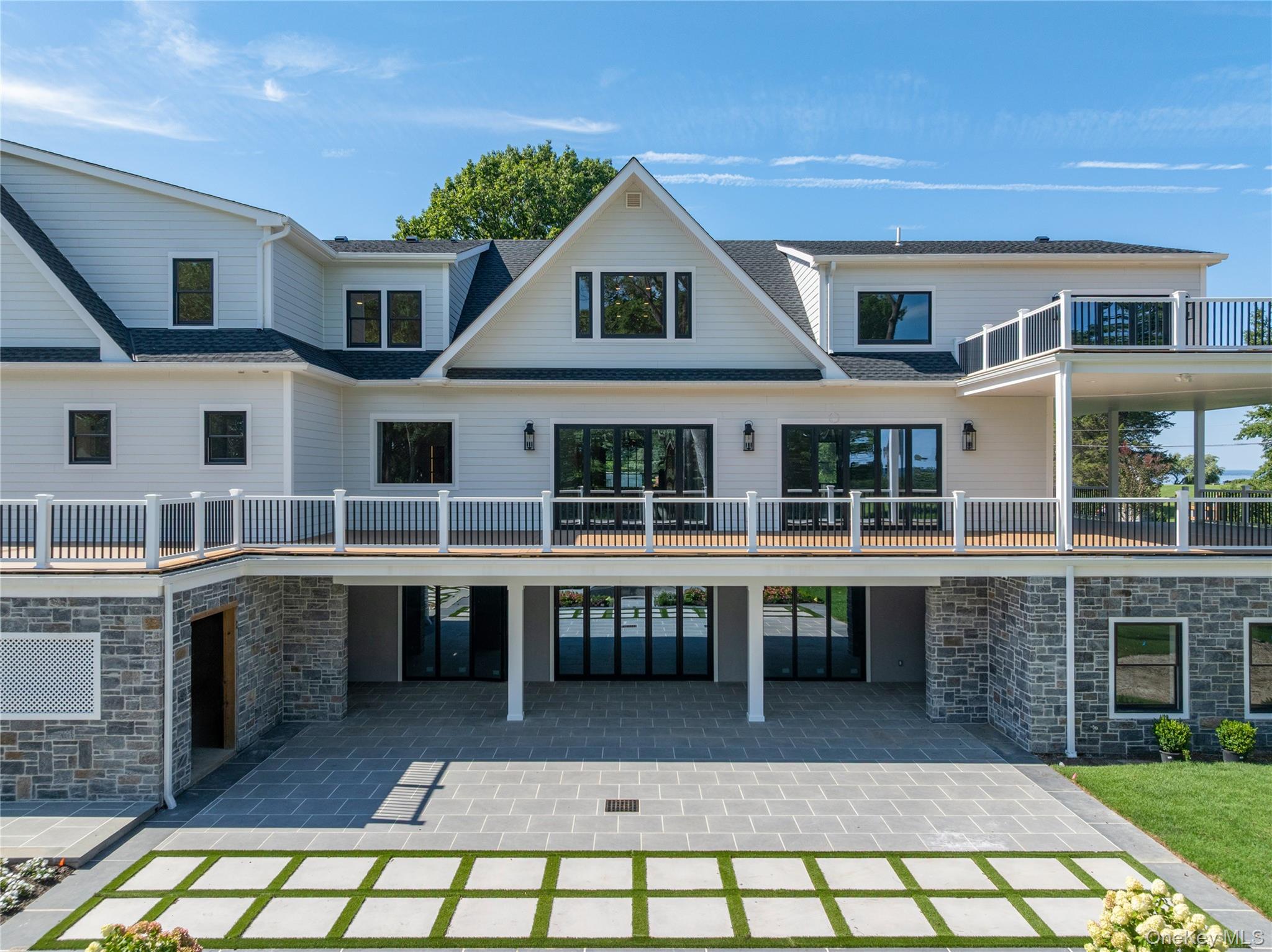 Large white home with a raised deck, covered stone patio, and geometric paver driveway.