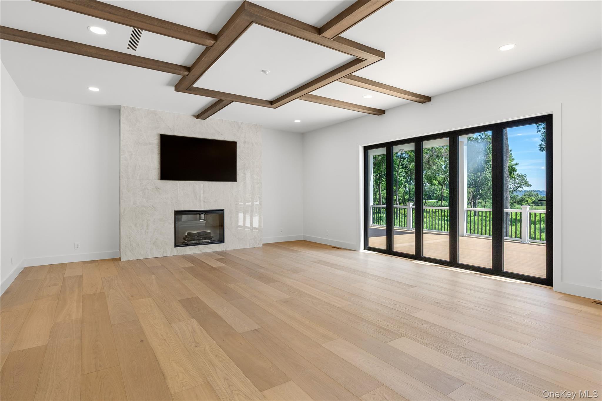 Vacant living room with a marble fireplace, geometric wood ceiling beams, and sliding doors to a leafy deck.