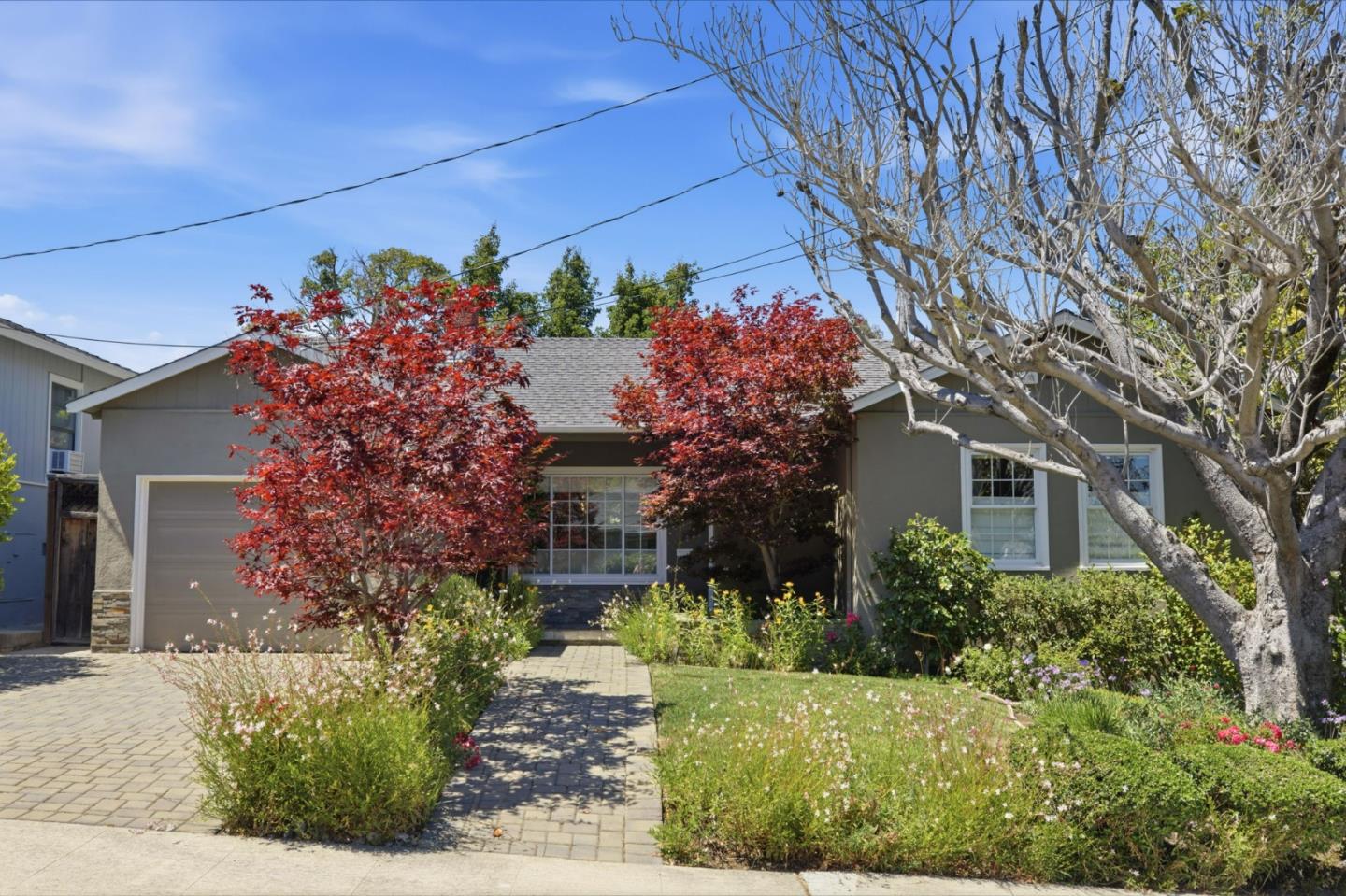 Gray single-story house with an attached garage, paved walkway, and red-leaved trees in the front garden.