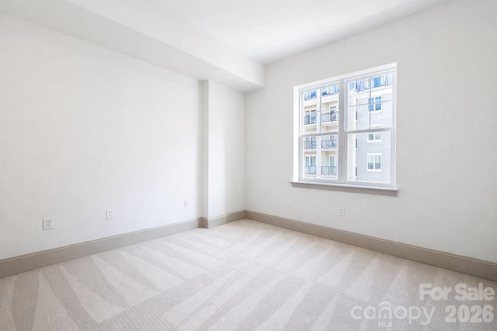 Empty carpeted room with white walls and a large window overlooking neighboring buildings.