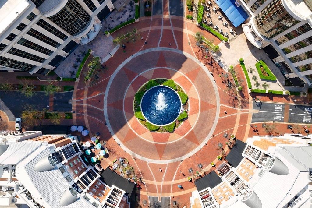 Aerial view of a circular plaza centered on a fountain, surrounded by buildings and outdoor seating.