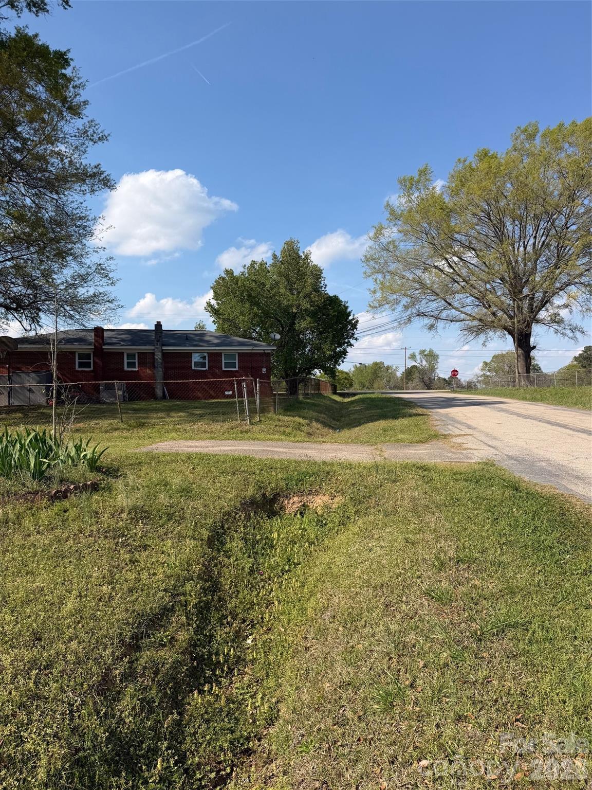Grassy roadside lot with a drainage ditch, chain-link fence, and brick house beside the road.