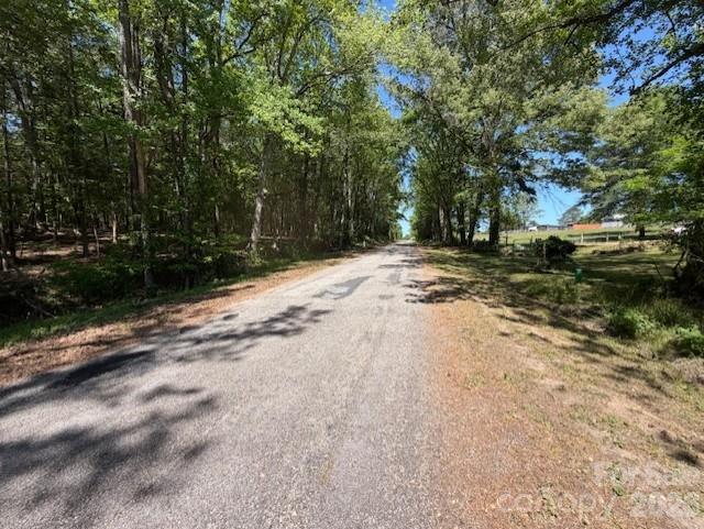 Paved road bordered by mature trees, with a grassy area and fence on the right.