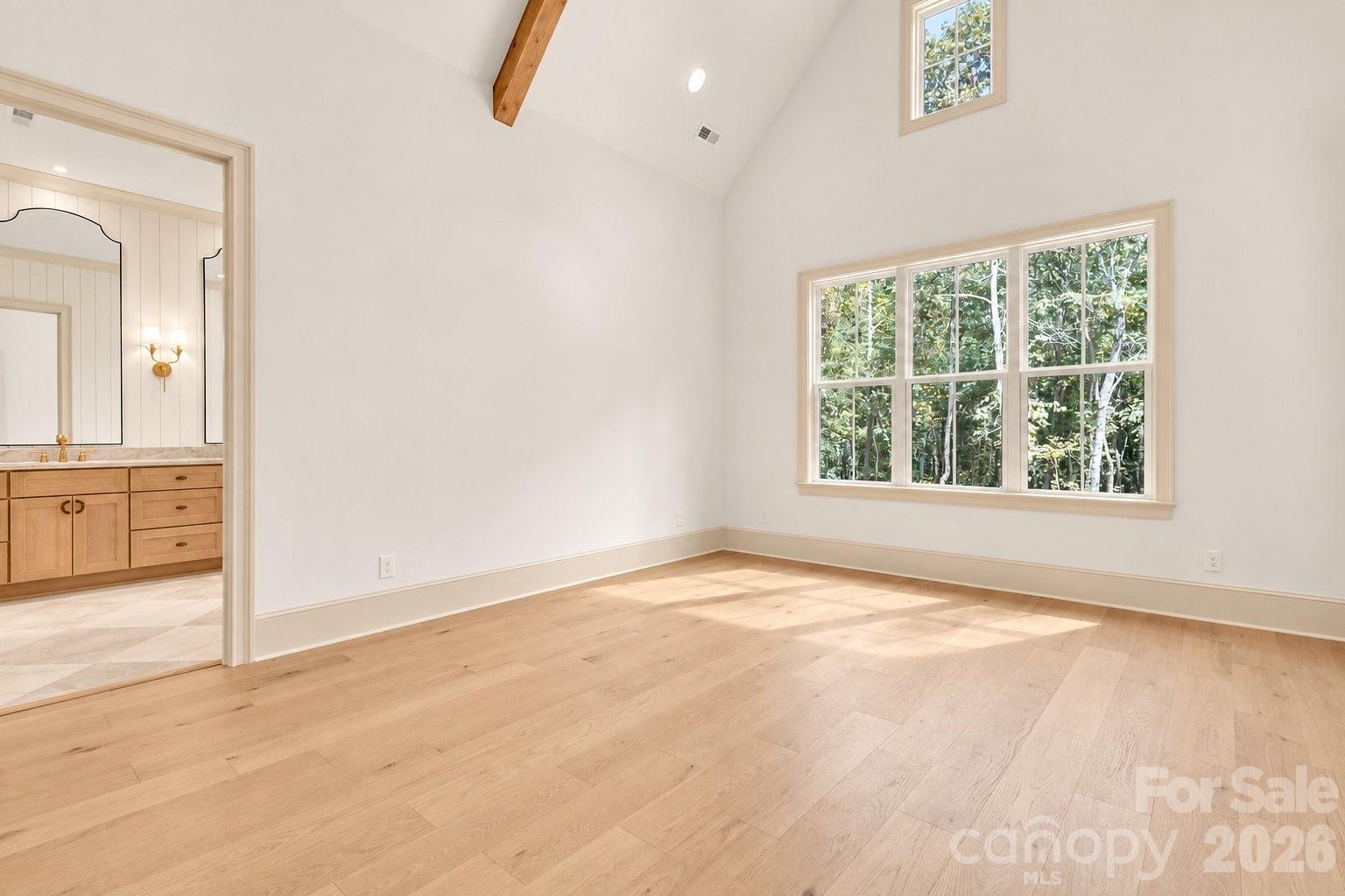 Empty bedroom with vaulted ceiling, pale hardwood floors, and a large window overlooking trees.