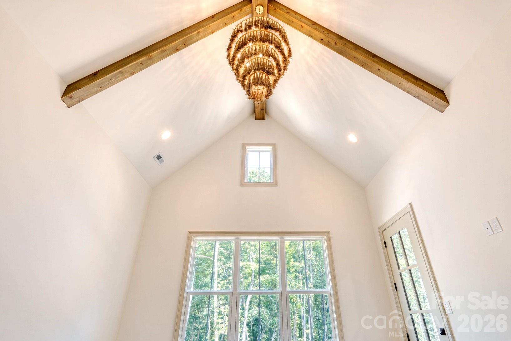 Vaulted room with exposed wooden beams, a tiered chandelier, and large windows overlooking trees.