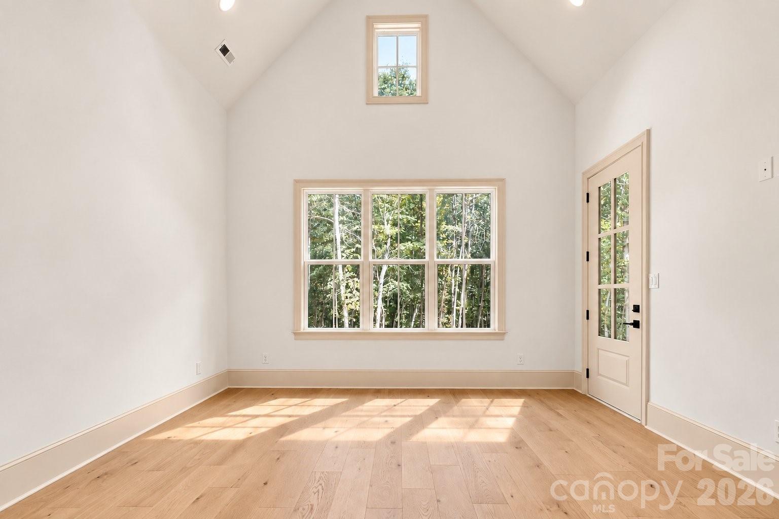 Bright vacant room with vaulted ceiling, light wood floors, large window, and glazed exterior door.