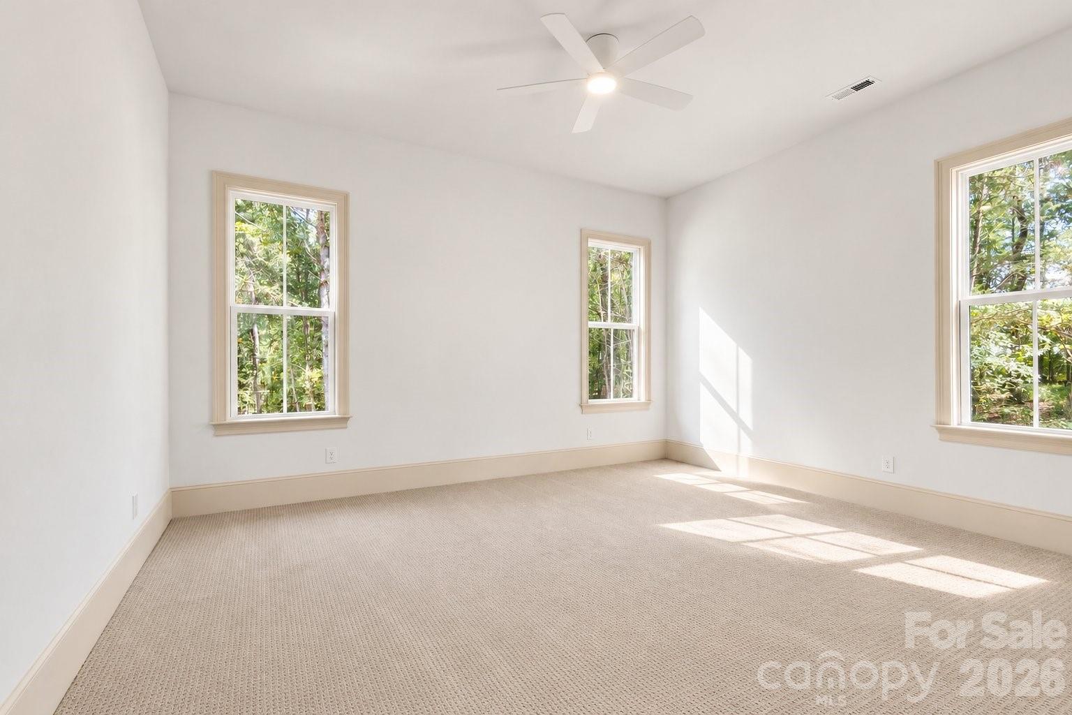 Empty carpeted bedroom with three tree-facing windows and a ceiling fan.