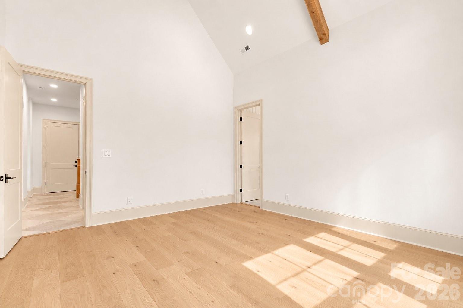 Vacant room with light hardwood floors, vaulted ceiling, exposed wood beam, and sunlight across the floor.