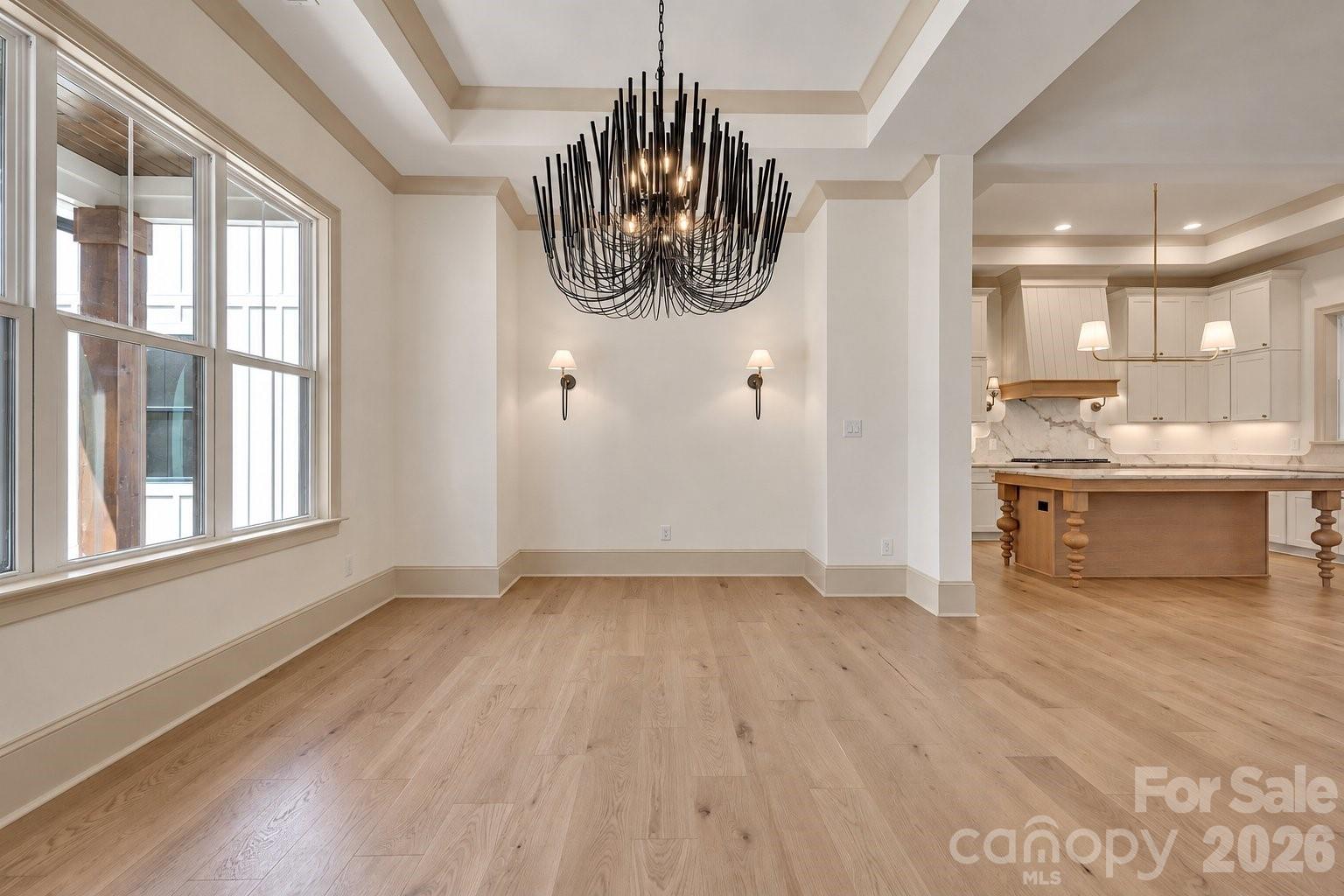 Empty dining room with a large black chandelier, tall windows, and an adjoining white kitchen.