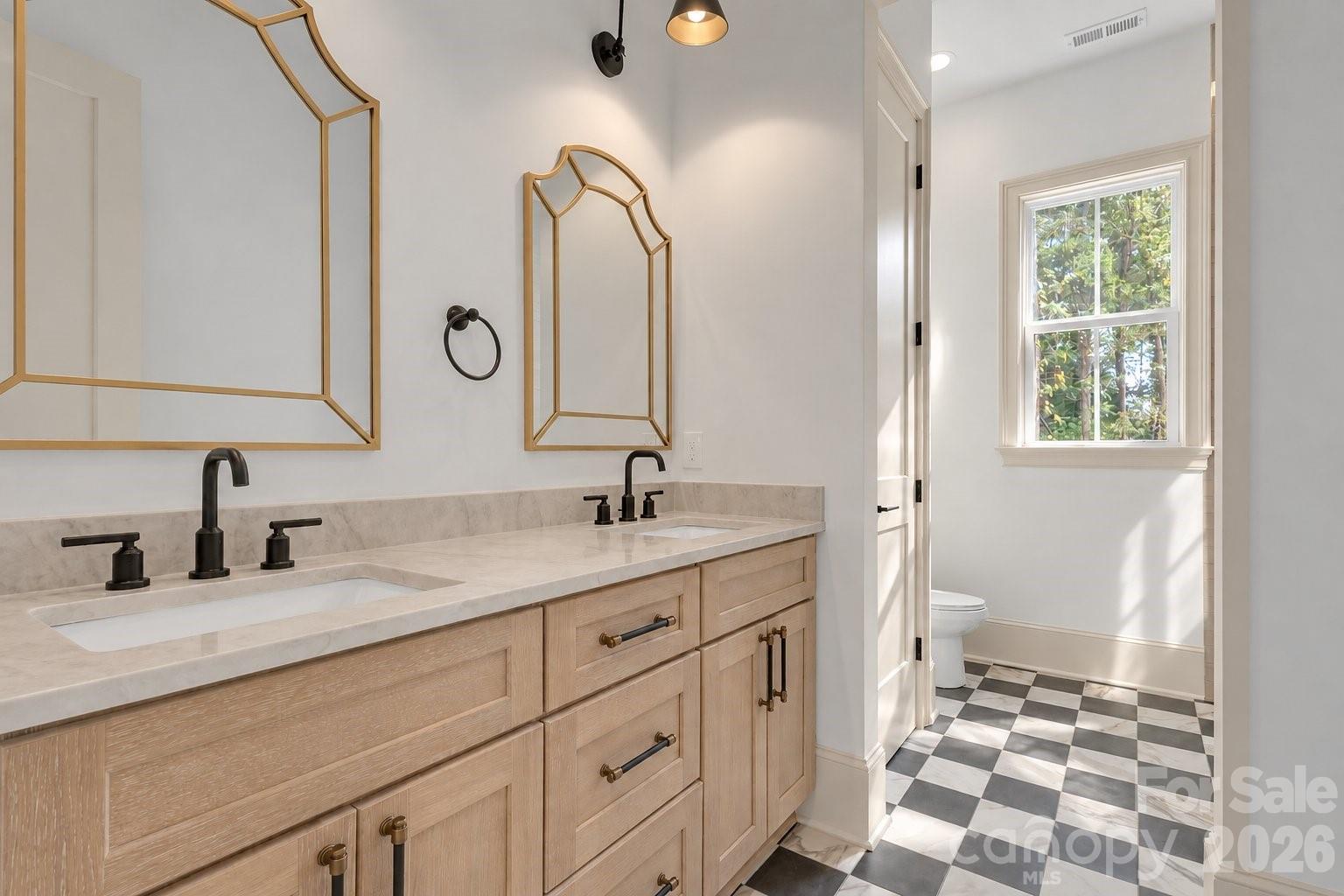 Bathroom with a double-sink vanity, gold-framed mirrors, and adjoining toilet area with checkerboard floor.