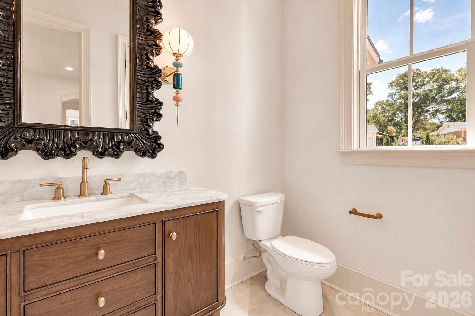 Bathroom with marble-topped wood vanity, ornate mirror, brass fixtures, and a large window.