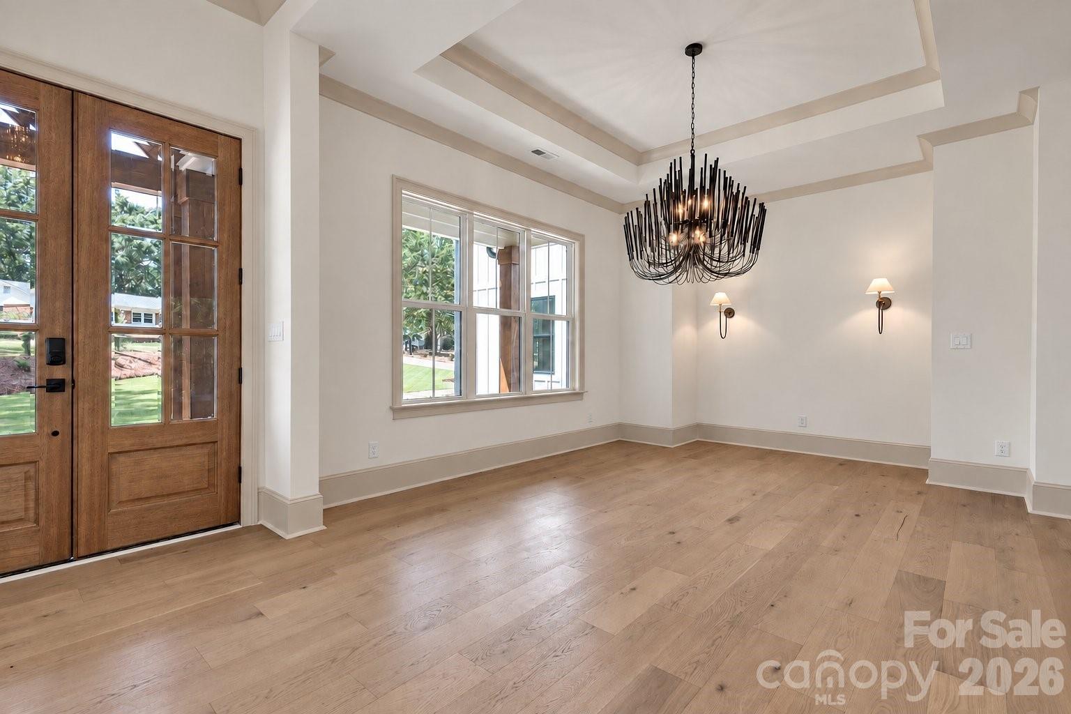 Empty dining room with a sculptural black chandelier, tray ceiling, and glass-paneled wooden door.
