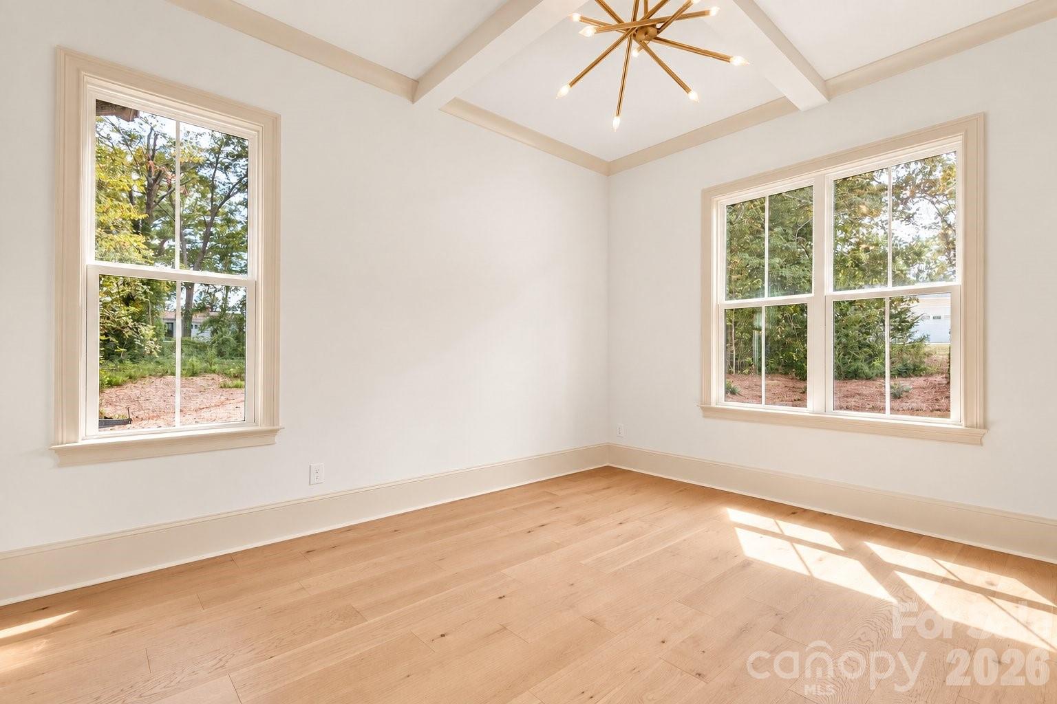 Empty room with pale hardwood floors, large windows, and a vaulted tray ceiling with a gold light fixture.