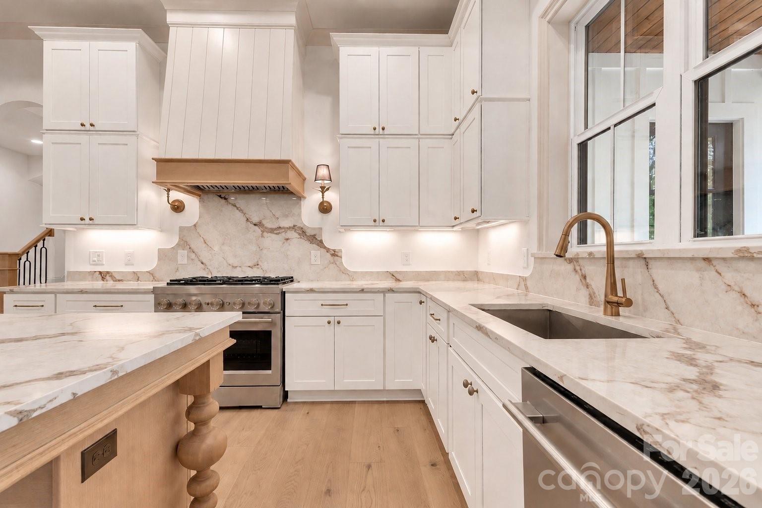 Kitchen with veined marble counters and backsplash, white cabinetry, brass fixtures, and large windows.