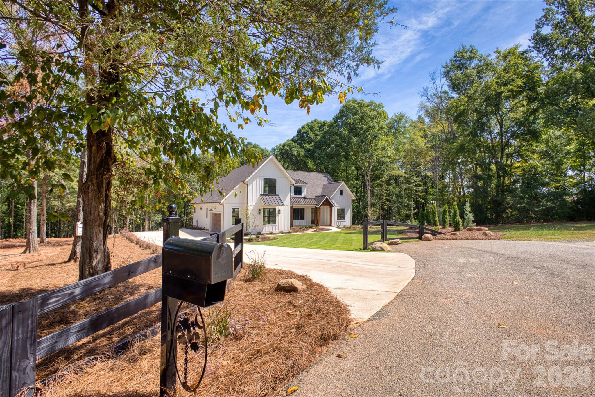 White farmhouse-style home among trees, with a curved driveway, black mailbox, and split-rail fence.