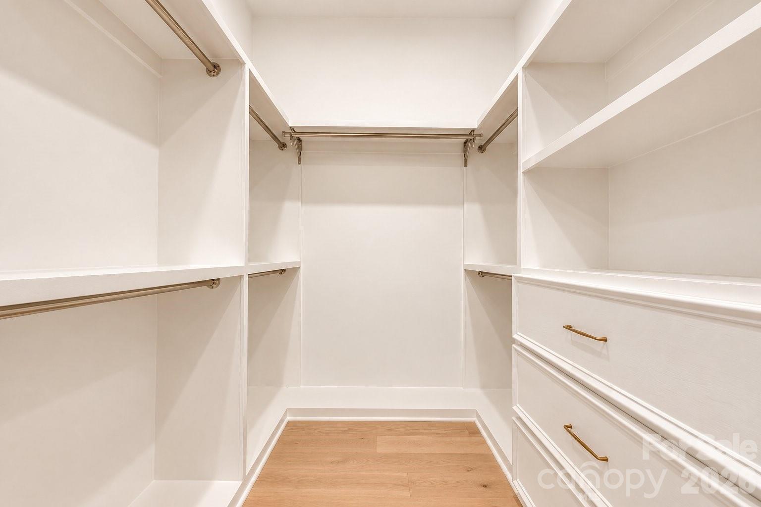 Walk-in closet with white built-in drawers, brass-colored hanging rods, and light wood flooring.