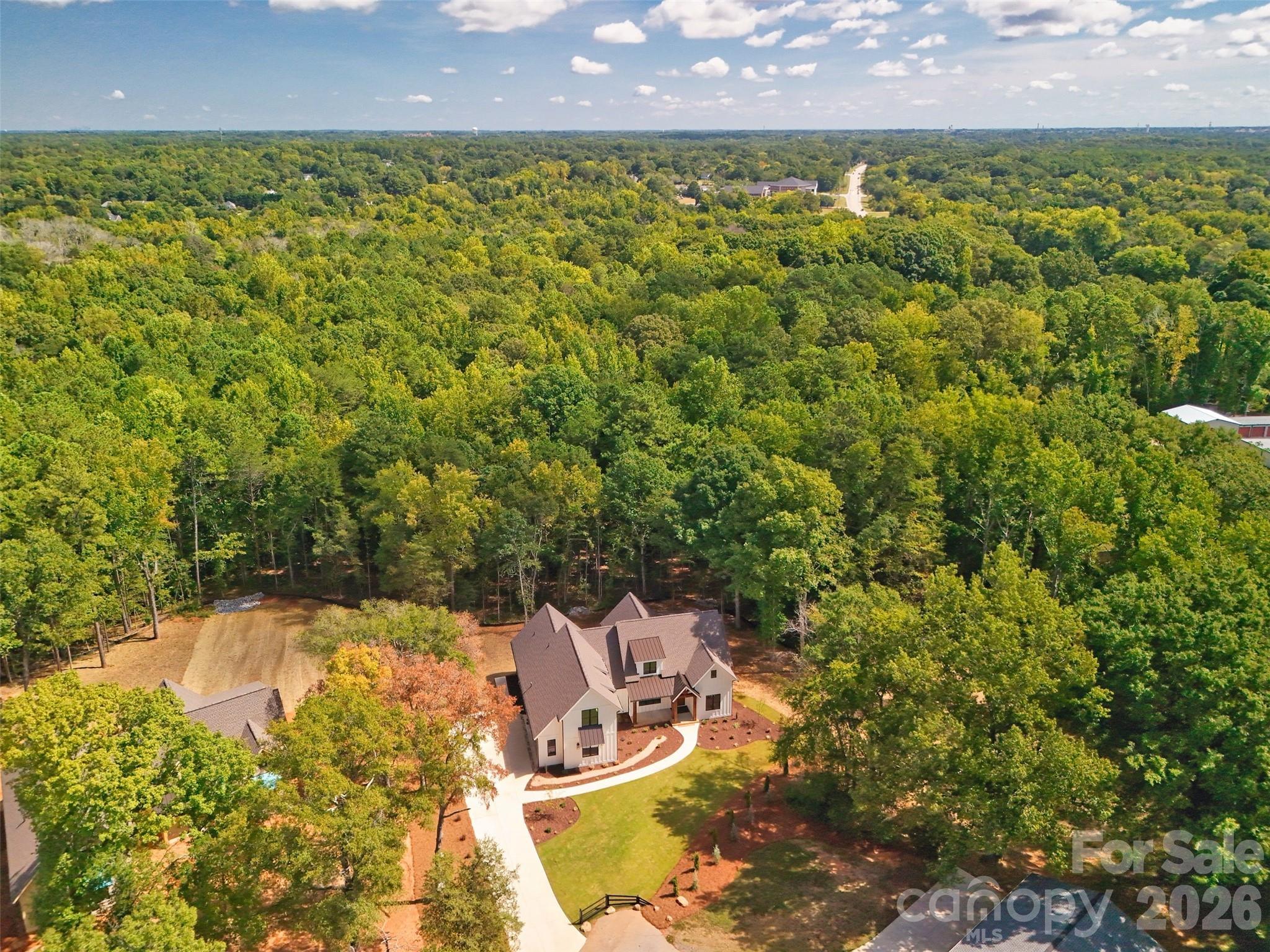 Aerial view of a house amid dense woods, with a curved driveway and landscaped lawn.