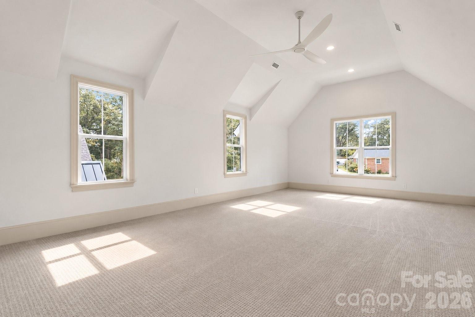 Vacant carpeted room with a vaulted ceiling, ceiling fan, and three sunlit windows.