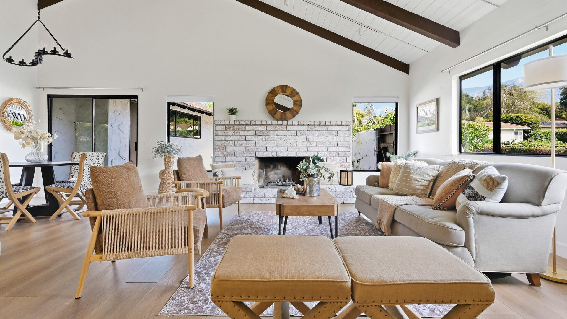 Living room with a white brick fireplace, vaulted beam ceiling, and large windows overlooking greenery and mountains.