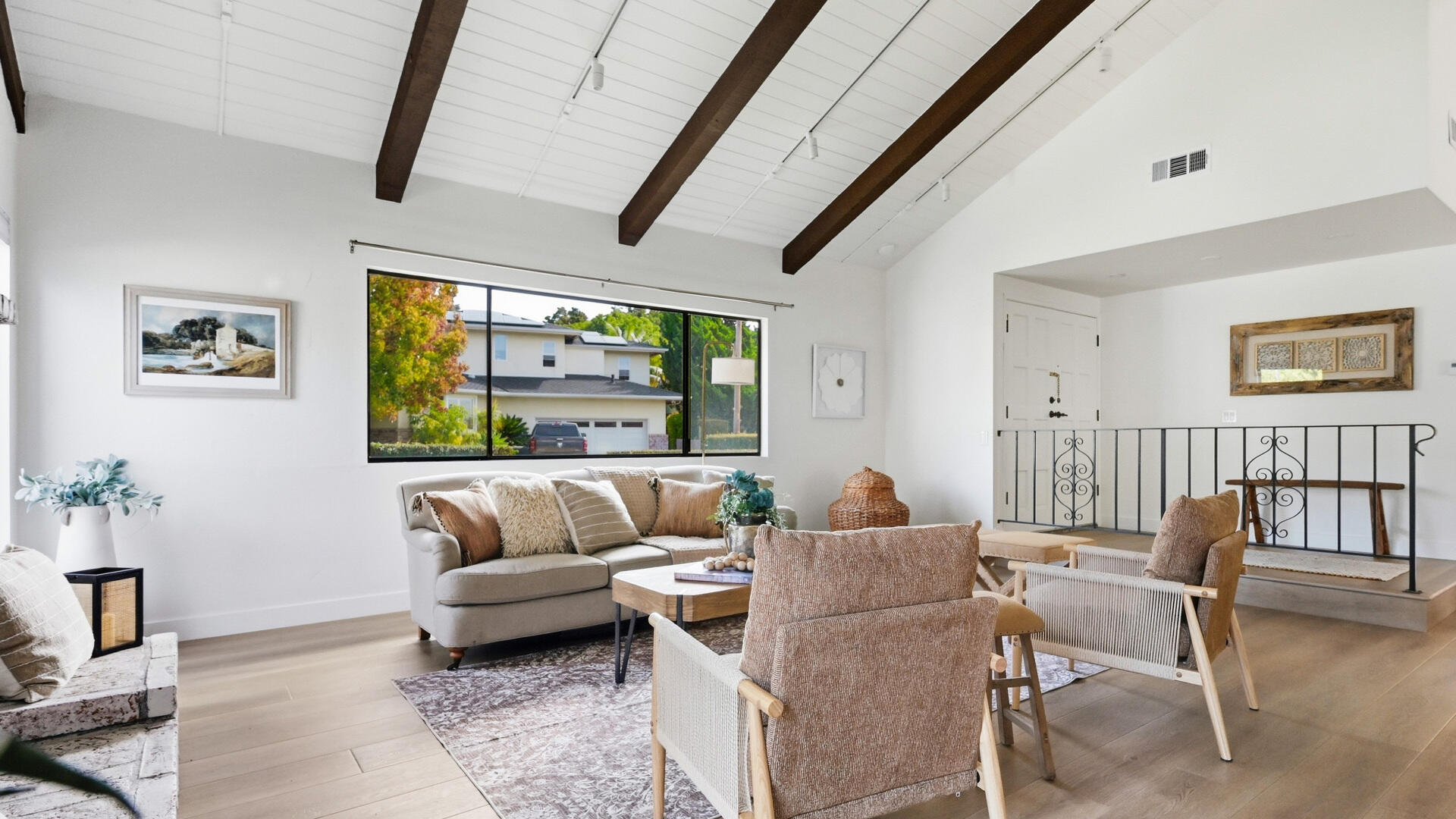 Bright living room with exposed beams, a wide picture window, neutral seating, and a decorative metal railing.