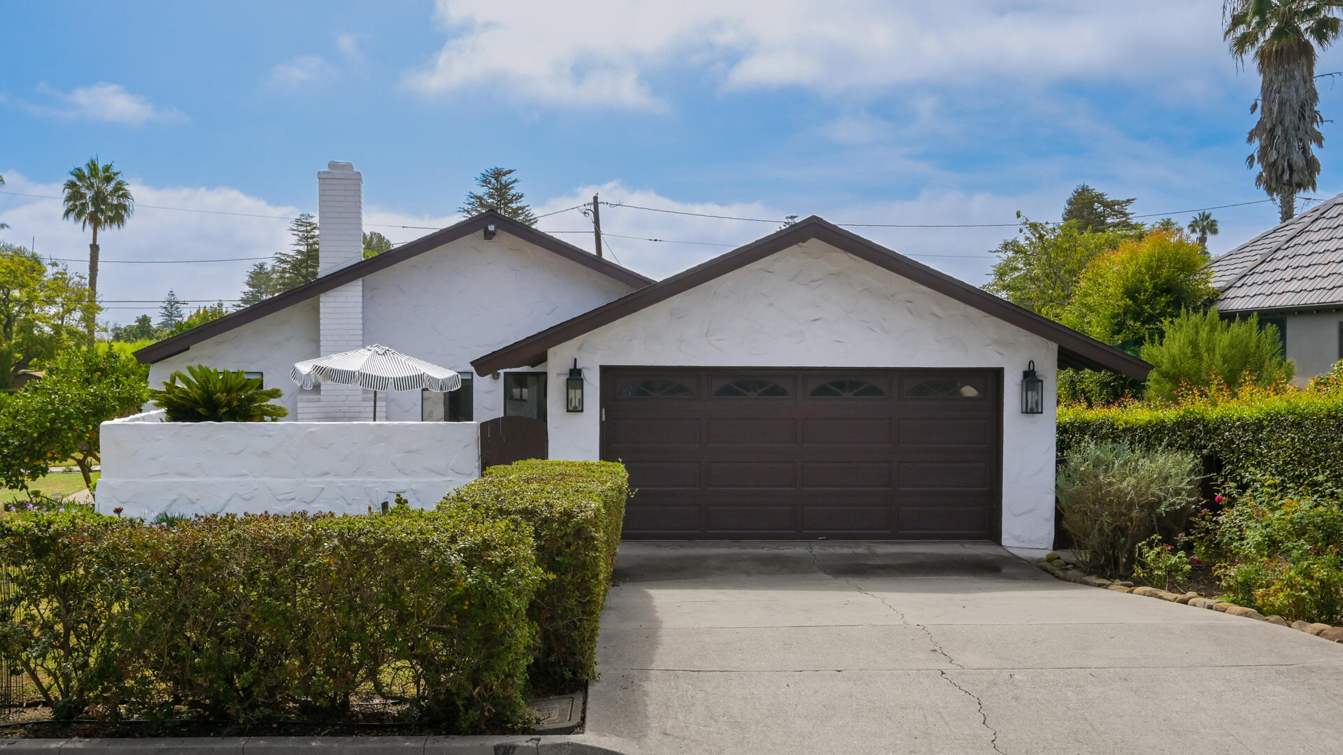 Single-story white stucco home with a dark double garage door, front hedges, and a striped patio umbrella.