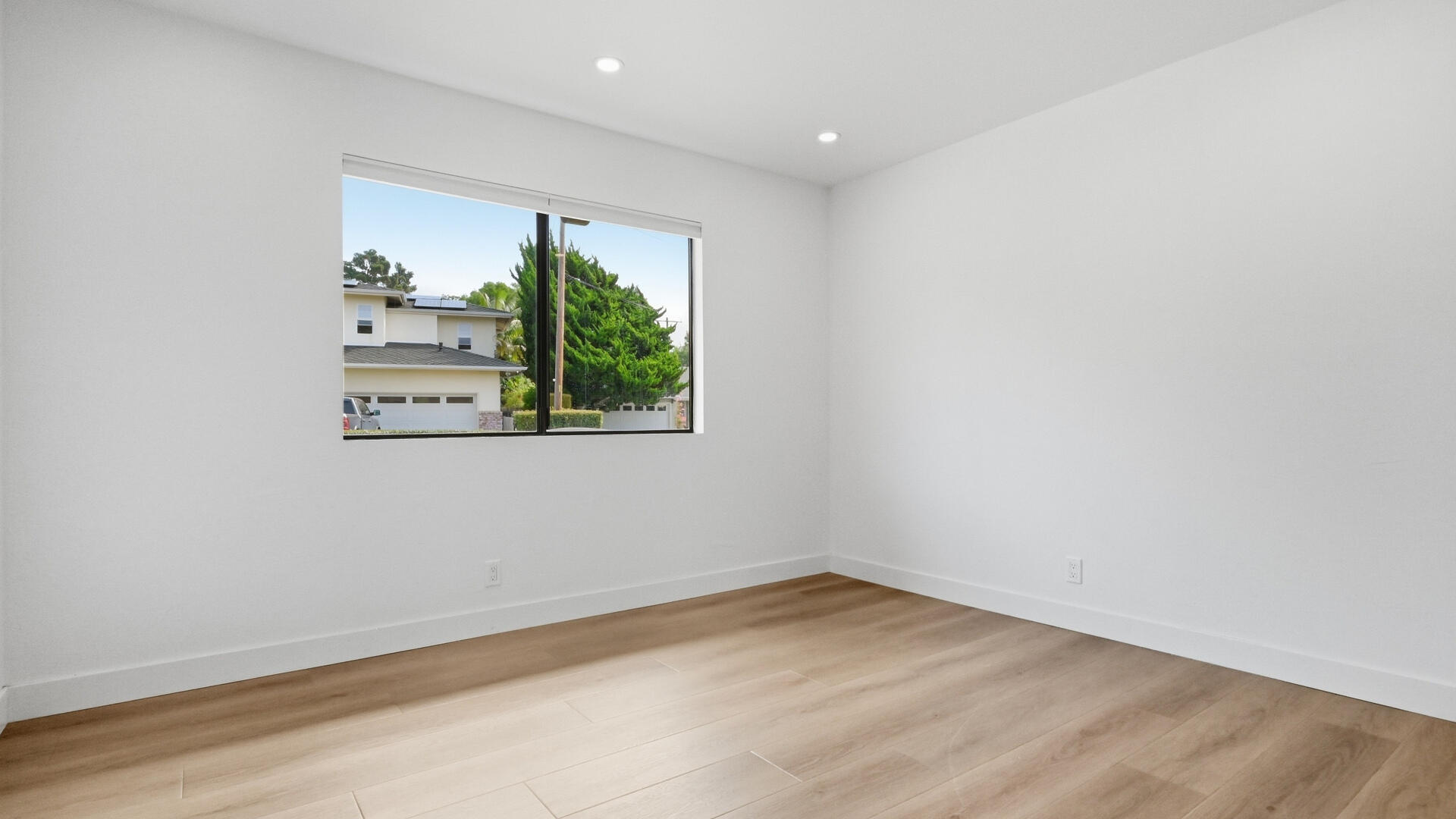 Empty room with light wood flooring and a wide window overlooking trees and a neighboring home.