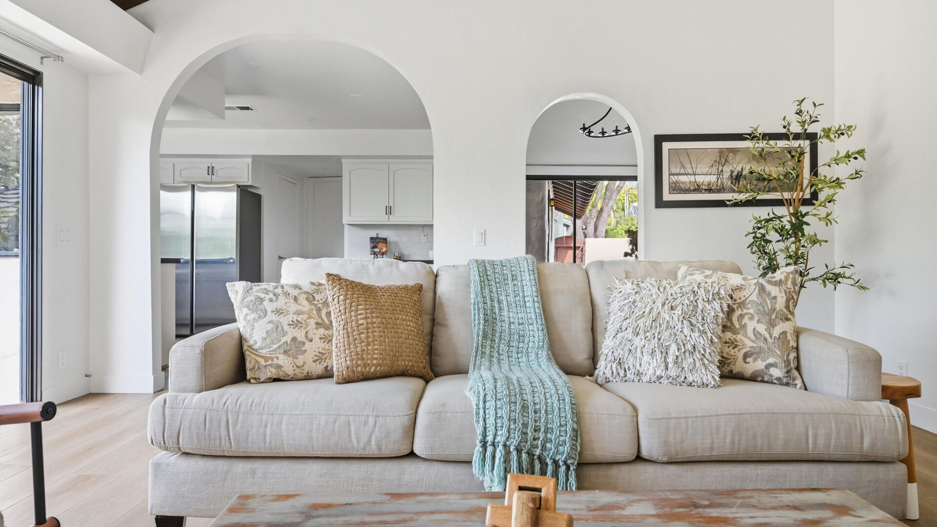 Living room with cream sofa and blue throw, framed by arches opening to a white kitchen.