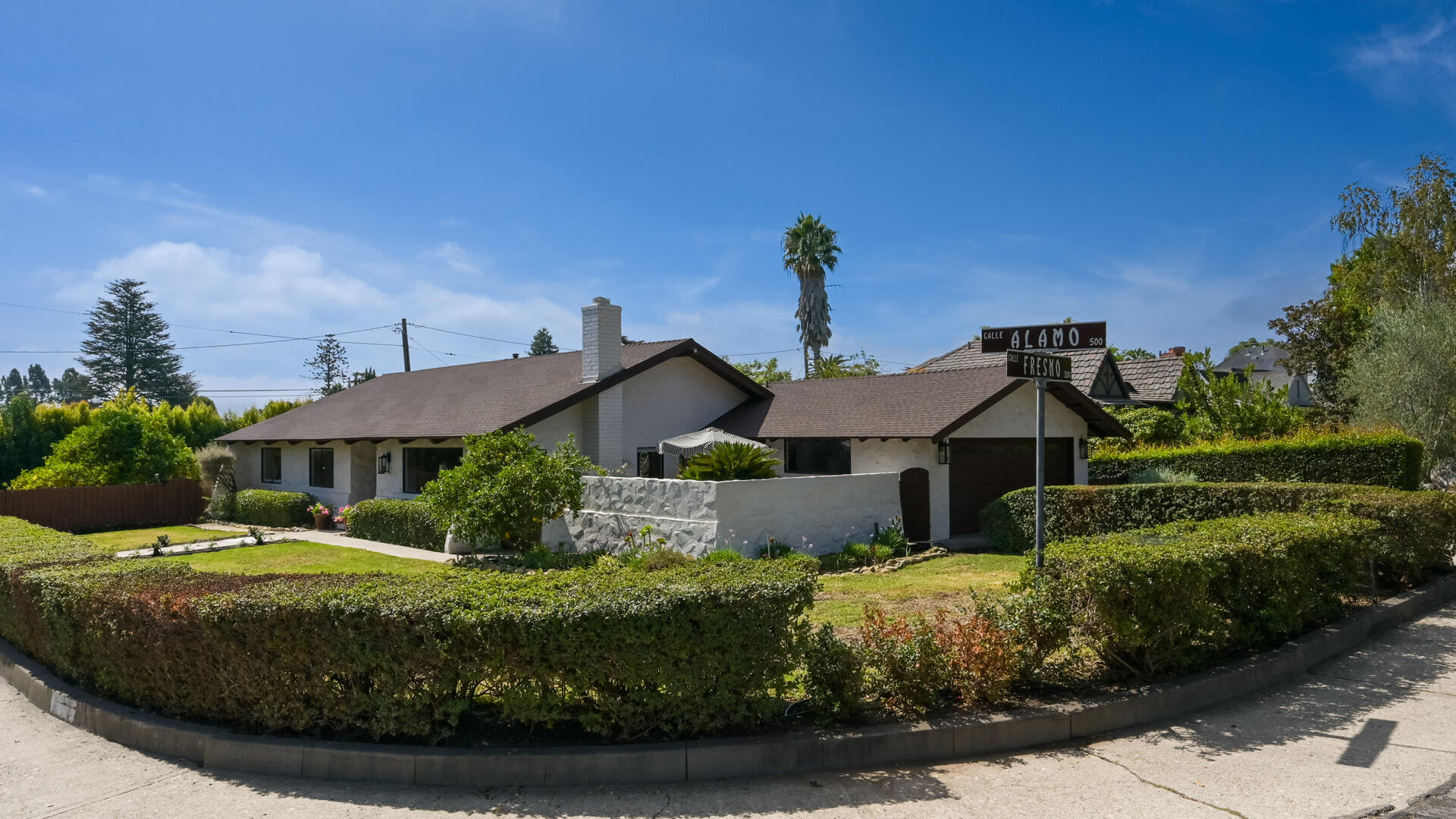 Single-story white stucco house with a brown roof, landscaped hedges, and a Calle Alamo street sign.