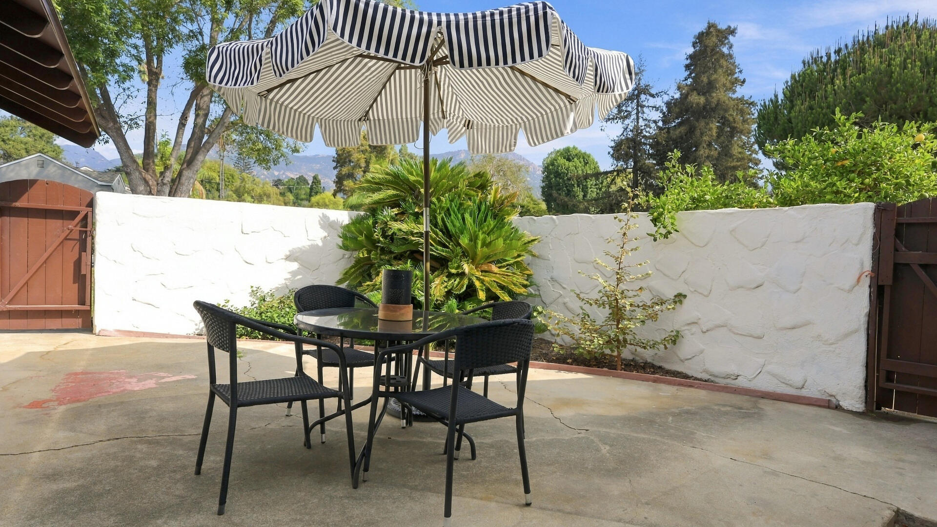 Patio seating beneath a striped umbrella, with a white wall and distant mountains.