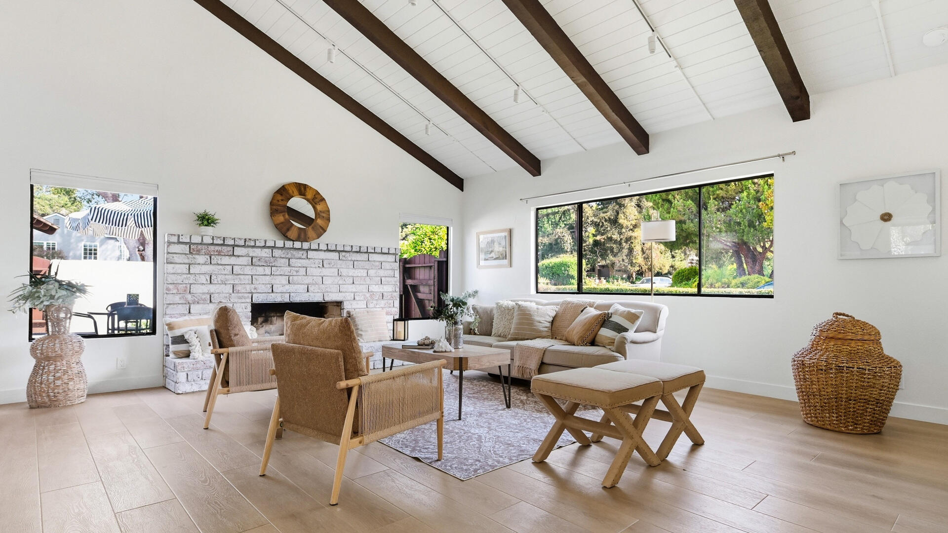 Staged living room with a white brick fireplace, vaulted beamed ceiling, and wide windows overlooking greenery.