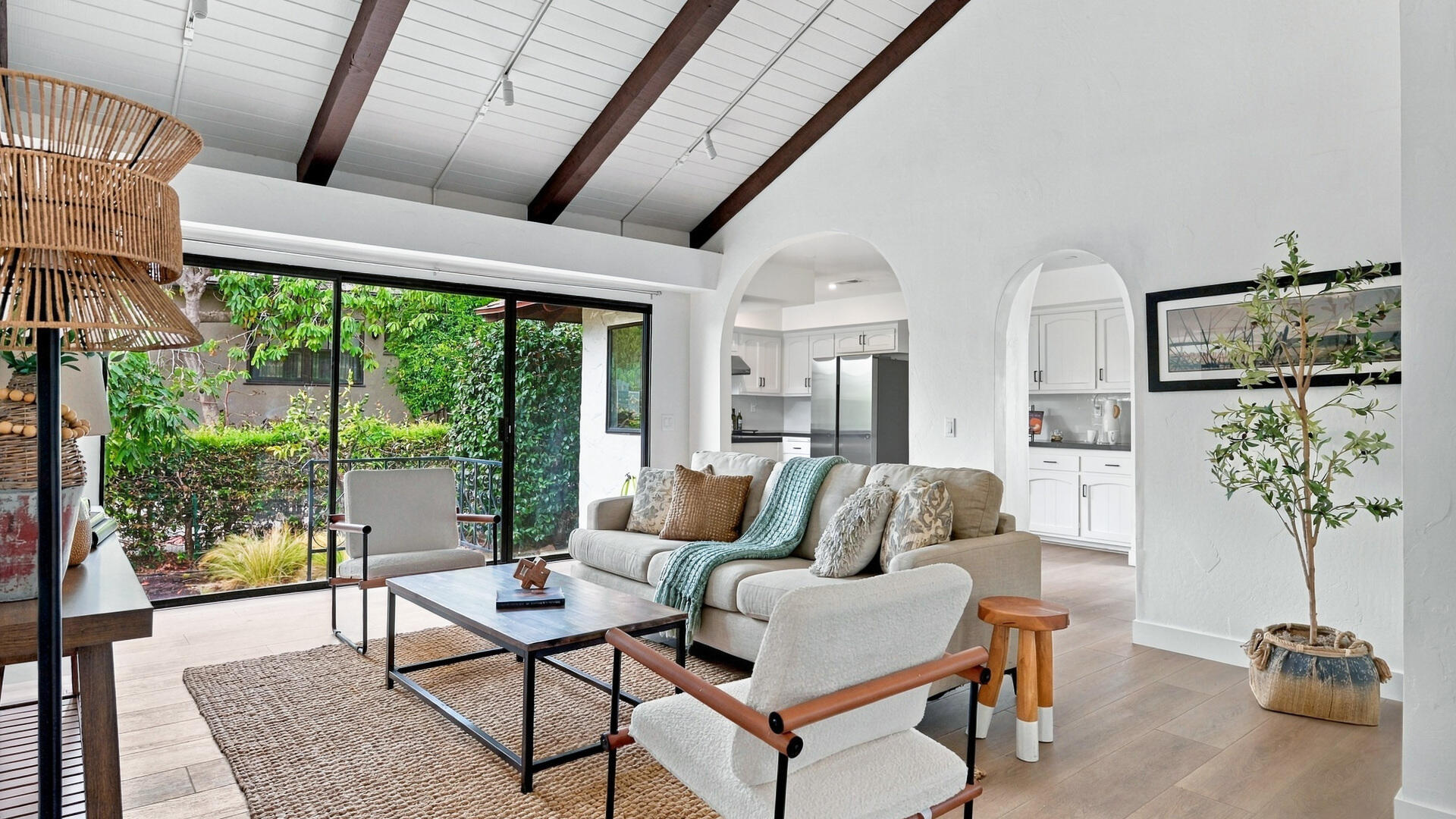 Living room with exposed ceiling beams, arched kitchen openings, and sliding doors overlooking a garden.