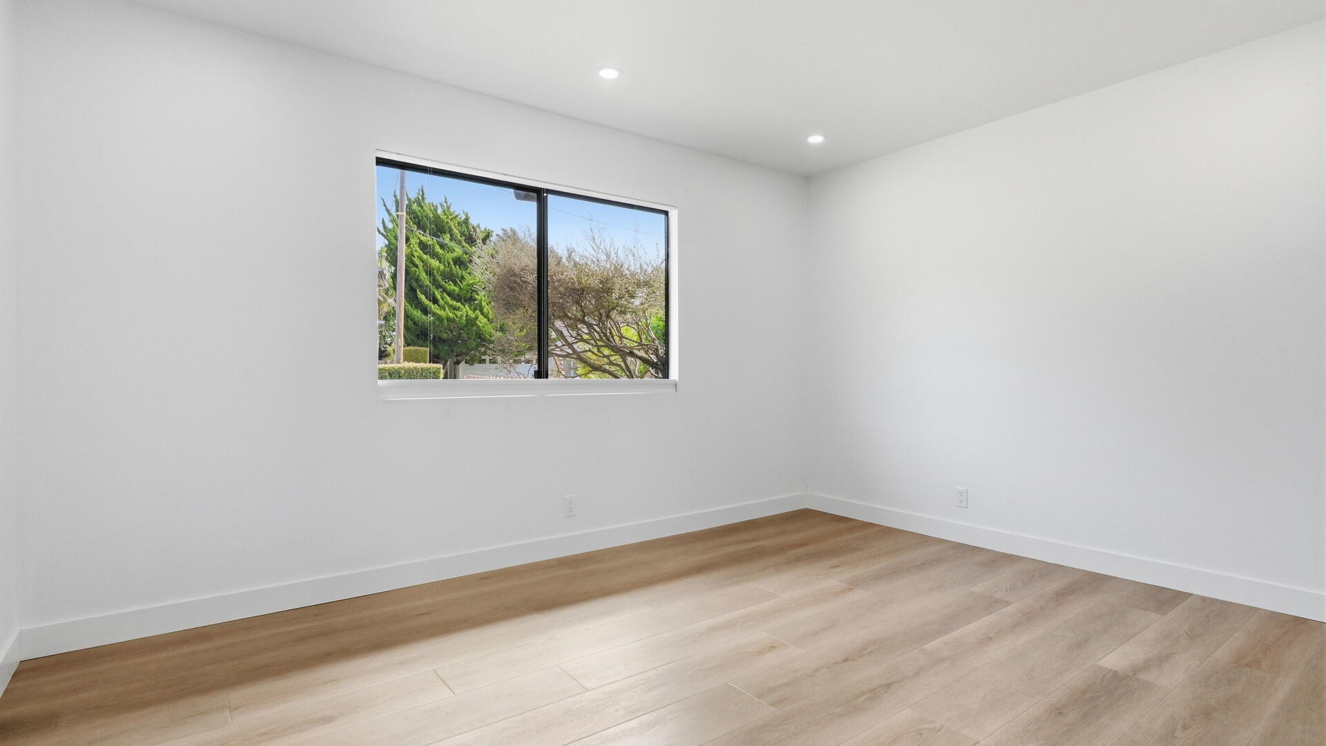 Empty room with light wood floors, white walls, and a large window overlooking trees.