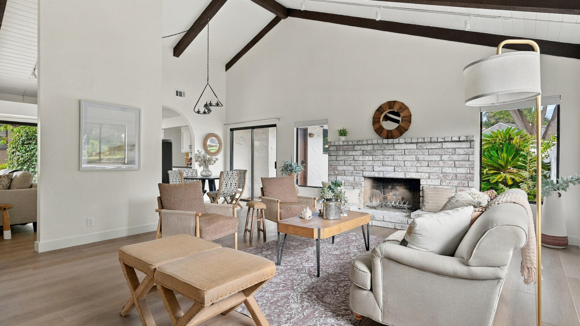 Furnished living room with exposed beams, a white brick fireplace, and large windows overlooking greenery.