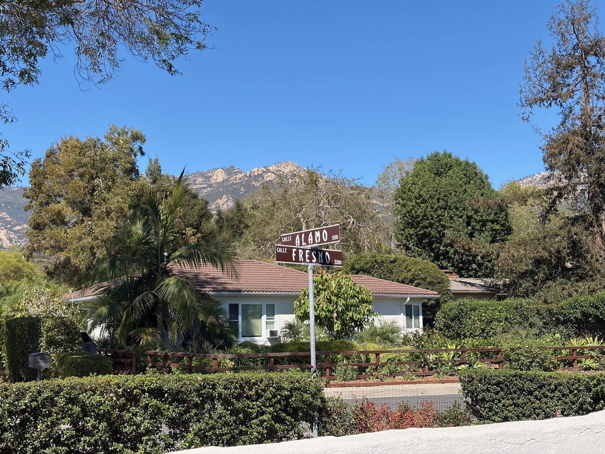 Single-story home behind leafy landscaping, with Calle Alamo and Calle Fresno signs and mountains beyond.