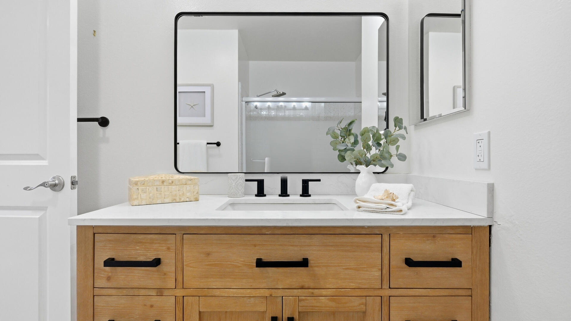 Bathroom vanity with a wood cabinet, white countertop, black-framed mirror, and glass shower.