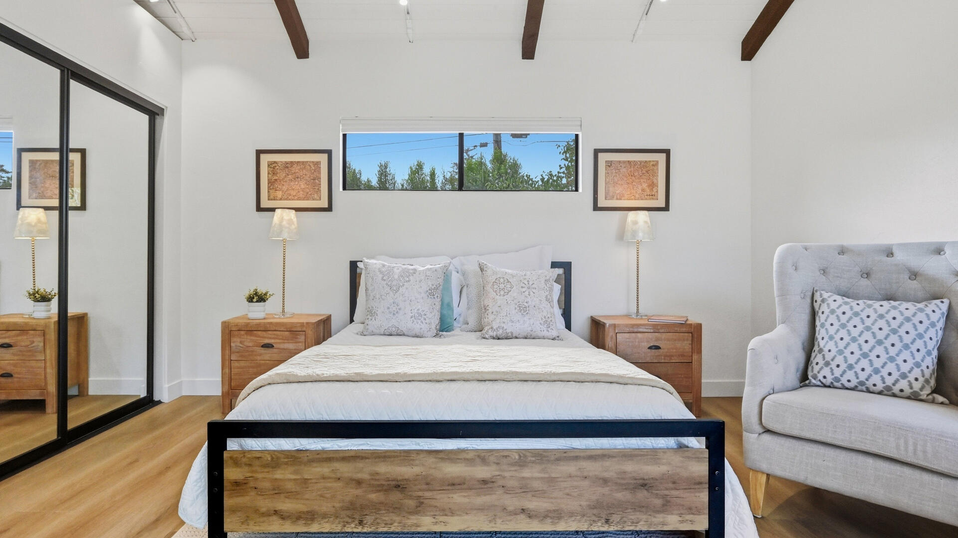 Staged bedroom with a wood-and-black bed, mirrored closet doors, exposed ceiling beams, and a gray armchair.